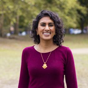 Headshot of Sheena Sood. Sheena is wearing a purple shirt and a gold necklace while standing in a park.