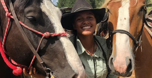 Jasmine Coates '22 an african american woman wearing a hat standing with two horses.