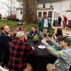 A group of older and younger alumni are seated at a round table outside having lunch.