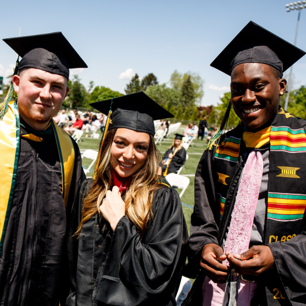 Three Students with cap and gown looking into the camera on the field at commencement.
