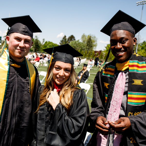 Three Students with cap and gown looking into the camera on the field at commencement. 