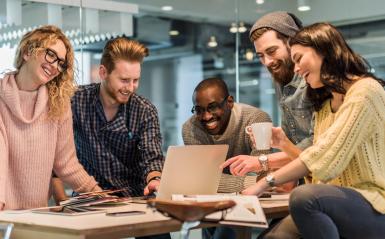 Young professionals gathered around a high-top table talking, laughing and looking a phones and a computer.