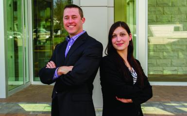 Professional looking 30-something man and woman standing back to back and smiling at camera.