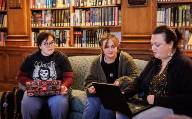 Two students and a professor are working on their own laptops and having a discussion.