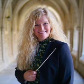 Headshot of Lauren Ryals standing in a cathedral-like outdoor hallway. Lauren is middle aged with long curly blonde hair.