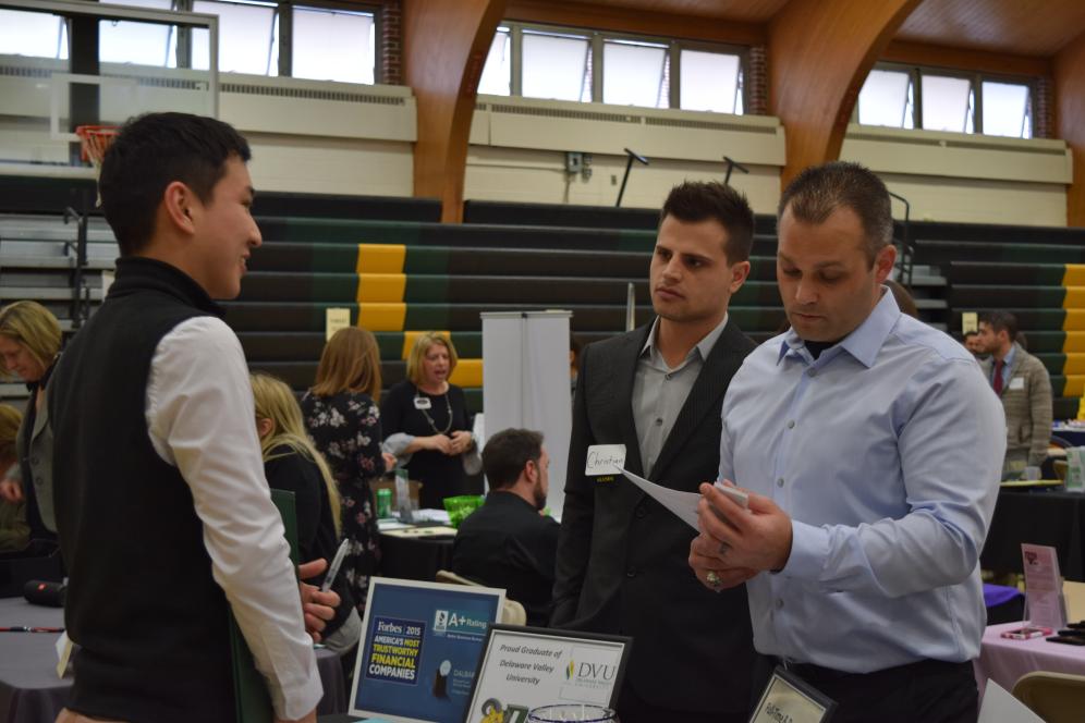 A student speaks with prospective employers at an information table at Delaware Valley University's Job and Internship Fair while an employer representative looks over his resume.