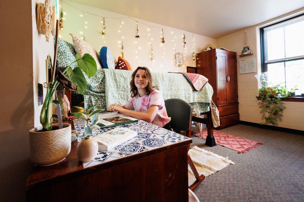 A female student is reading a book at her desk in her dorm room.