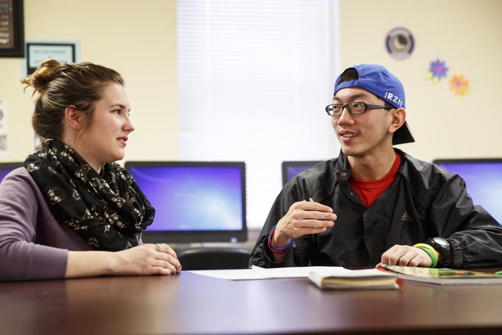 A male and female student are seated at a table talking about a paper at the writing center.