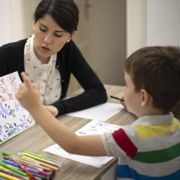 A woman is working with a child at a table with paper and crayons.