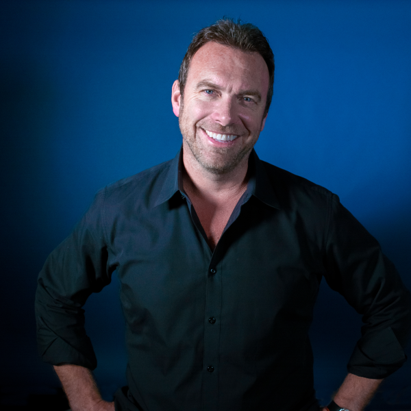 A headshot of Alex Boylan smiling with a blue collared shirt in front of a blue background.