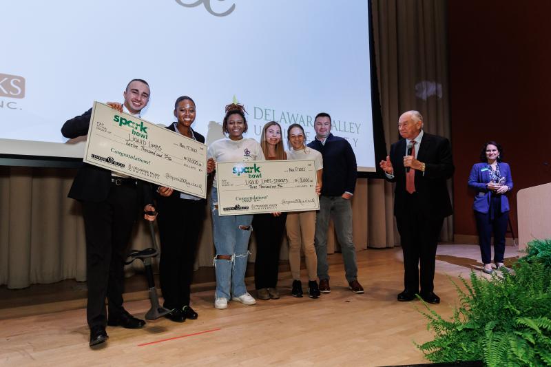 Liquid Limbs and their student business consultants line up for a photo holding giant checks. 