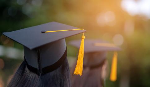 Graduates sit in file. We see their caps.